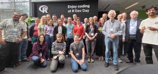 About two dozen R Dev Day participants gathered for a group photo in a bright, glass-walled atrium, in front of a sign reading 'Enjoy coding at R Dev Day'.